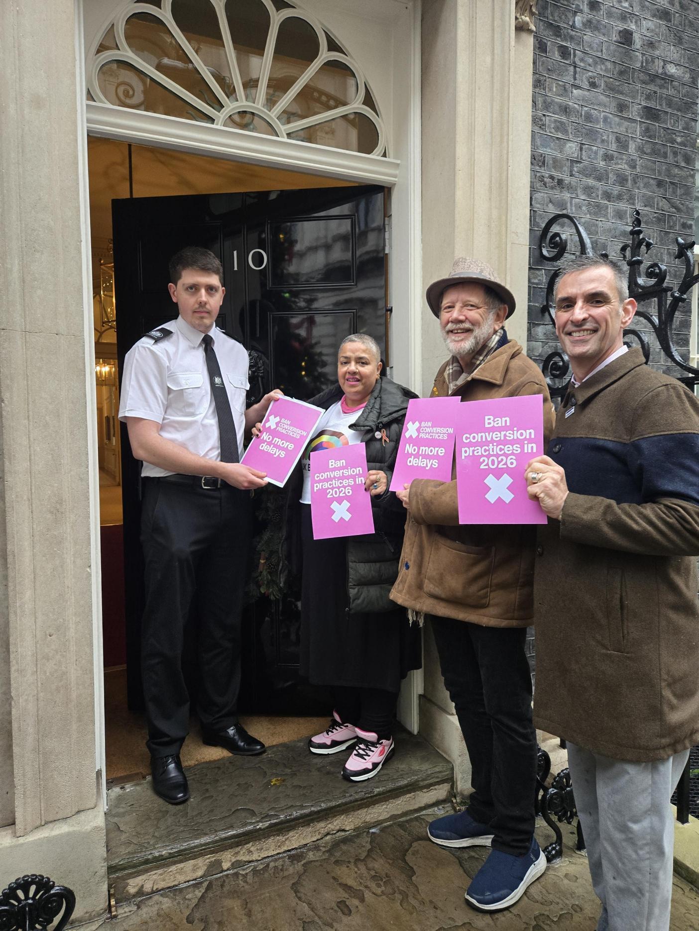 Three people stand outside 10 Downing Street delivering a letter, holding signs that read “Ban conversion practices in 2026.”