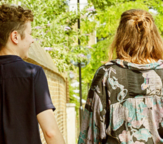 Two people with their backs to us walking along the pavement