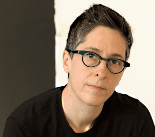 A person with dark glasses and short black hair in a black top, against a black and white background.