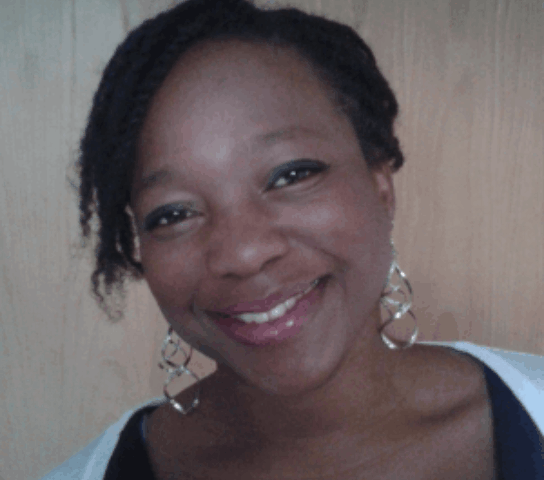 A woman in a light green top with gold earrings against a wooden wall