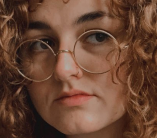 A person in round-frame glasses with long wavy brown hair.