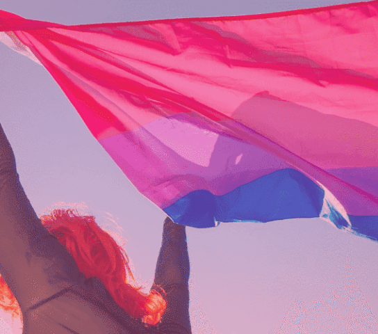 Person with orange hair holding the bi flag against a blue sky.