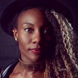 A Black woman looks at the camera. She has wavy blond hair and wears a black hat. She has a gold-disc earring, and wears a band around her neck. The left side of her face is in shadow.