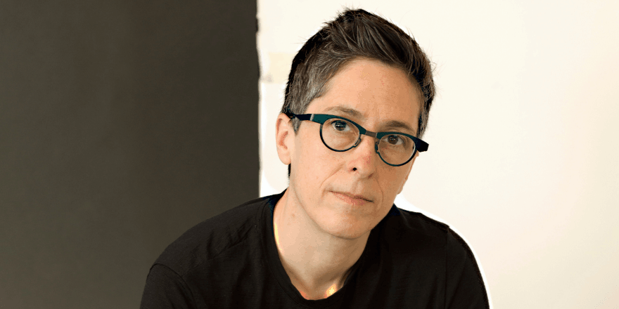 A person with dark glasses and short black hair in a black top, against a black and white background.