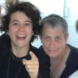 Two white women, one younger, one older, look at the camera, which is looking down on them. The younger woman points her finger at it and is smiling.