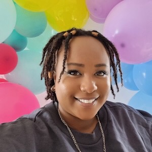 A Black woman smiles at the camera. She has short hair, wears a grey jumper and a necklace. Behind her are multicoloured balloons.