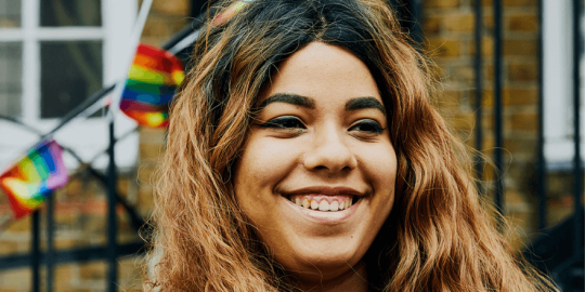 A Black woman in a leather jacket with long hair stands against a brick wall, smiling. There are rainbow flags in the background.
