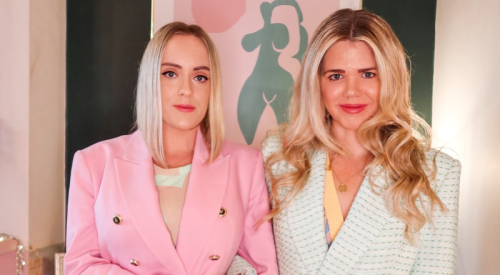 Two white women, one in a pink jacket, the other in a white jacket with green checks, stand in a mostly pink room and look at the camera. They both have long blonde hair.
