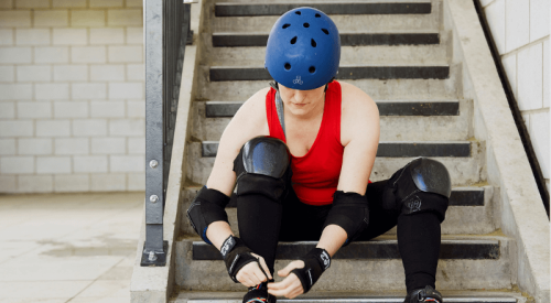 A person in a red top, black leggings and a blue helmet, laces up rollerblades while sitting on concrete steps