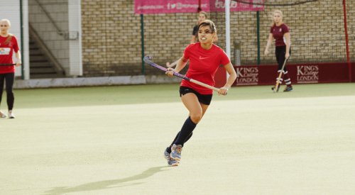A person in a red top, black shorts, wearing Rainbow Laces, places hockey on astroturf