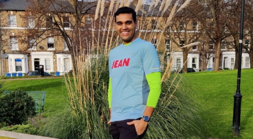 A man of colour stands smiling at the camera, wearing running clothes, in a green open space in front of a row of houses