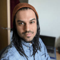 Person of colour with dreadlocks, in a blue shirt with a red beanie hat, looks at the camera from an inside room 