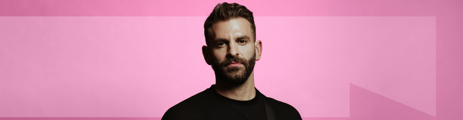 White person stands in front of a pink background, wearing black, with a beard and short hair. An arrow is visible in the background