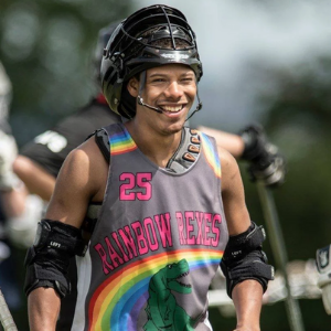 Person of colour in lacrosse outfit that reads Rainbow Rexes smiles in a field