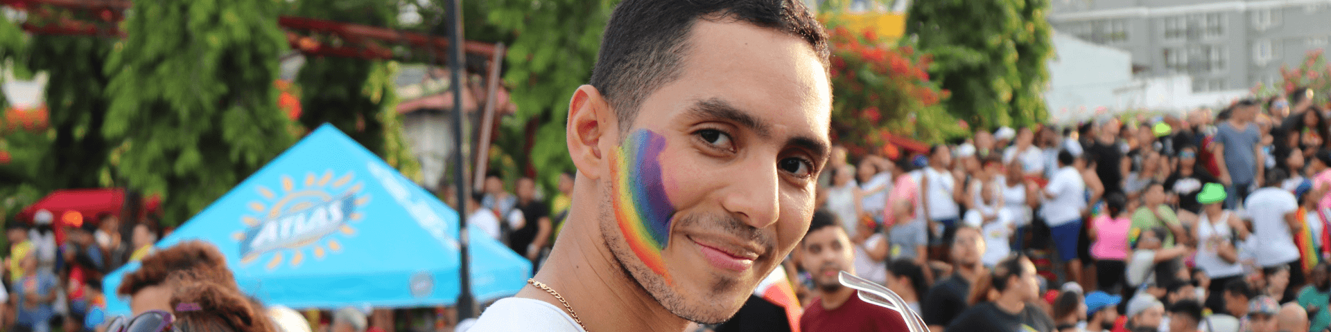 Person in a white t-shirt with a Pride flag painted on their cheek looks sideways at the camera. They're outside, at a festival