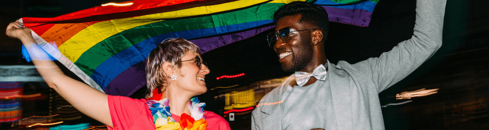 A Black person wearing a bowtie, and a white person wearing a garland of flowers, hold a Pride flag up and smile at each other. They're in a city at night, and the lights behind them are blurred.